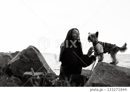 Black and white photo of a young woman with long hair gently touching her small Yorkshire terrier doggy in clothes on seaside rocks. Emotional portrait showing love connection of human and lovable pet 133271844