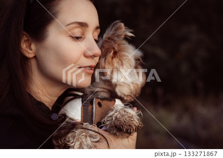 Woman lovingly embraces her small dog outdoors. A tender moment of affection. A girl is holding Yorkshire terrier doggy, enjoying a peaceful moment. Woman lovingly embraces her small dog outdoors. A tender moment of affection. A girl is holding Yorkshire terrier doggy, enjoying a peaceful moment. 133271867