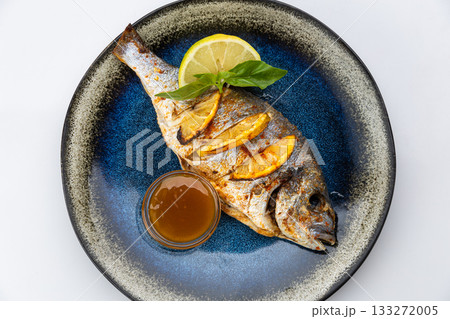 Whole grilled dorado with lemon, basil and sauce on a blue ceramic plate, white background 133272005