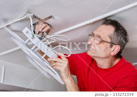 Electrician connects wiring of new LED chandelier to ceiling mount in rural house, fixture supported by hand, installation process continuing. 133272354