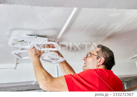 Installation process shows electrician mounting modern LED chandelier onto ceiling in rural house using both hands during connection of lighting fixture. 133272367