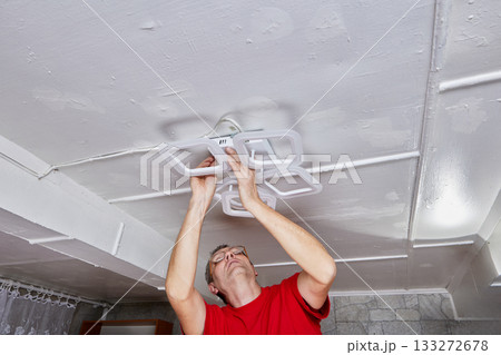 LED chandelier is positioned on ceiling while electrician performs installation in rural house connecting wires and securing fixture. LED chandelier is positioned on ceiling while electrician performs installation in rural house connecting wires and securing fixture. 133272678