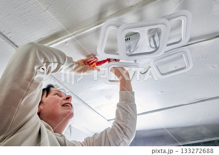 Attaching LED chandelier to ceiling in rural house performed by female installer, geometric fixture and hand tool visible, installation phase ongoing. Attaching LED chandelier to ceiling in rural house performed by female installer, geometric fixture and hand tool visible, installation phase ongoing. 133272688