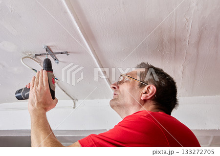 Fixing electrician uses cordless screwdriver to secure ceiling bracket for LED chandelier in rural house, wires held close to mounting area. Fixing electrician uses cordless screwdriver to secure ceiling bracket for LED chandelier in rural house, wires held close to mounting area. 133272705