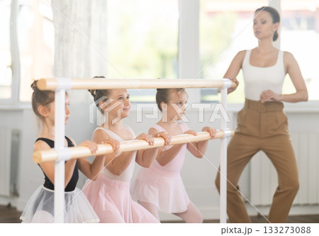 Teacher conducts class with children girls in ballet studio, observes doing grand plie exercise. 133273088