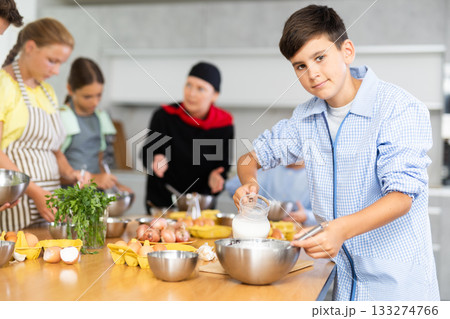 During cooking classes, teen boy carefully mixes ingredients in bowl with whisk During cooking classes, teen boy carefully mixes ingredients in bowl with whisk 133274766