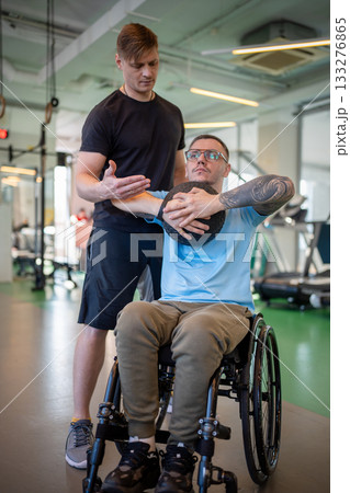 Man in wheelchair do upper body exercises with medicine ball under help of personal trainer in gym. 133276865