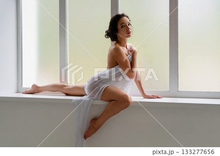 Woman in white fabric poses gracefully by large window in natural light 133277656