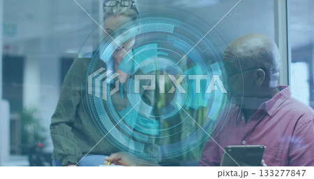 Woman wearing knit sweater and senior man reviewing data overlay at desk in office, holding tablet 133277847