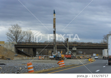 Workers are setting up drilling equipment next to highway under construction with partially built overpass overhead. Workers are setting up drilling equipment next to highway under construction with partially built overpass overhead. 133278717