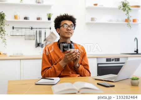 A young man in glasses holds a coffee cup while looking up thoughtfully in a bright kitchen setting. A young man in glasses holds a coffee cup while looking up thoughtfully in a bright kitchen setting. 133282464