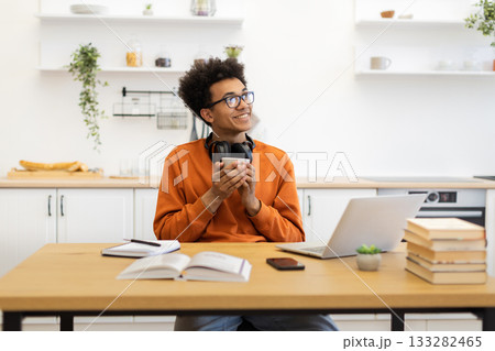 A young man with glasses smiles while holding a cup of coffee in his kitchen, with a laptop and books on the table. 133282465