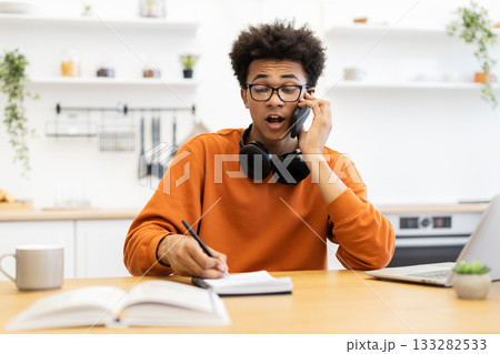 A young man with glasses is on a phone call while working at a wooden desk in a bright kitchen setting. A young man with glasses is on a phone call while working at a wooden desk in a bright kitchen setting. 133282533