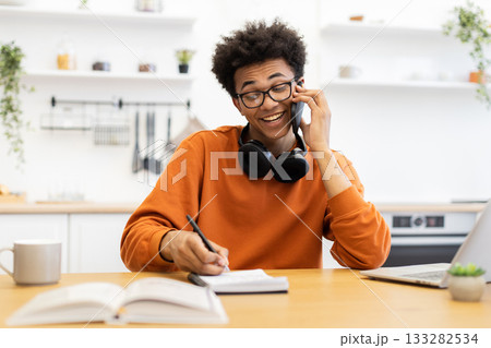 A young man with glasses smiles while talking on the phone and taking notes at his desk in a bright, modern kitchen. A young man with glasses smiles while talking on the phone and taking notes at his desk in a bright, modern kitchen. 133282534