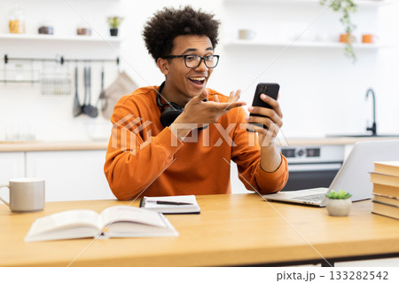 A young man with glasses is smiling and gesturing while looking at his phone, possibly on a video call, at home. 133282542