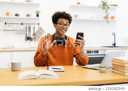 A young man with glasses waves during a video call, sitting at a table with a laptop and books in a bright kitchen setting. A young man with glasses waves during a video call, sitting at a table with a laptop and books in a bright kitchen setting. 133282543