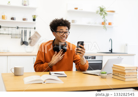A young man with glasses smiles while looking at his phone during a video call, sitting at a table in a bright kitchen. 133282545