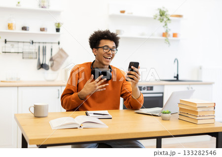 A young man laughs while on a video call, sitting at a table with a laptop and books in a bright kitchen setting. 133282546