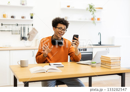 A young man with glasses waves during a video call while sitting at a table in his kitchen. A young man with glasses waves during a video call while sitting at a table in his kitchen. 133282547