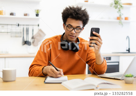 A young man with glasses is working at a table, using his phone and writing in a notebook. 133282558