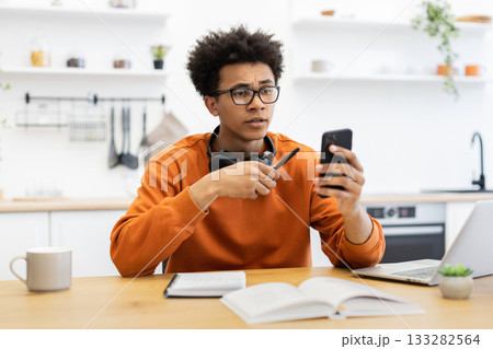 A young man with glasses looks at his phone with a concerned expression while sitting at a table in a kitchen. A young man with glasses looks at his phone with a concerned expression while sitting at a table in a kitchen. 133282564
