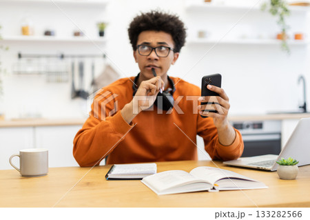 A young man in glasses contemplates while holding a phone and pen, with a book and laptop on the table. 133282566