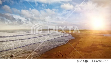Flat design showing curving shoreline, rolling waves on beach under sun and clouds over far dunes 133286262