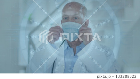 Senior doctor wearing coat adjusting surgical mask over face in hospital corridor, with tech screen Senior doctor wearing coat adjusting surgical mask over face in hospital corridor, with tech screen 133288322