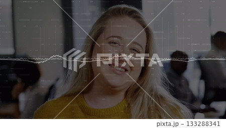 Smiling woman wearing mustard sweater sitting at office desk, monitors and audio waveform overlay Smiling woman wearing mustard sweater sitting at office desk, monitors and audio waveform overlay 133288341