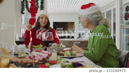 Sharing laughter mother and daughter wearing Santa hats and sweaters at dining table, with candle 133288473