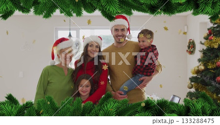 Smiling family group wearing Santa hats posing in living room, with golden snowflake confetti 133288475