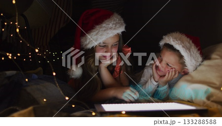 Lying siblings using glowing tablet in bedroom at home, with Santa hats and string lights 133288588