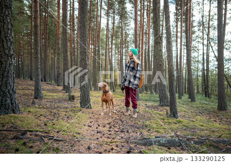 Woman and dog walk forest trail in autumn, both look upward as if spot bird in tree canopy Woman and dog walk forest trail in autumn, both look upward as if spot bird in tree canopy 133290125