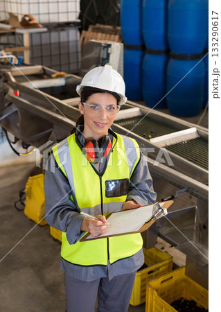 Female inspector inspecting conveyor in plant wearing helmet earmuffs glasses holding clipboard 133290617