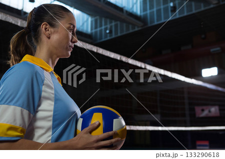Female volleyball player wearing jersey holding volleyball looking toward net in gym, copy space 133290618