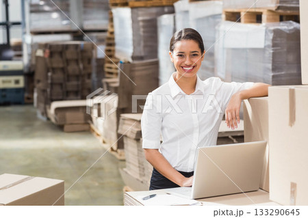 Woman in late twenties wearing white shirt checking laptop on boxes in warehouse, copy space 133290645