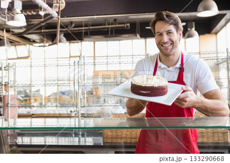 Male baker wearing red apron standing at bakery display counter holding frosted cake, copy space 133290668