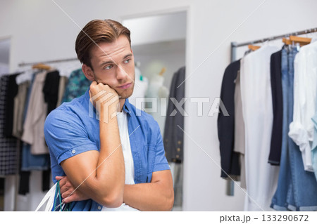 Man holding shopping bags, resting chin on hand and gazing at apparel racks inside clothing store 133290672