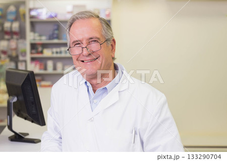 Senior male pharmacist in lab coat standing behind counter at pharmacy using computer monitor 133290704