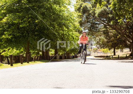 Thirtysomething woman riding bicycle along shady park path past wooden bollards wearing helmet 133290708