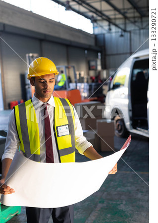 Asian man reviewing blueprint while wearing yellow hard hat and reflective vest in warehouse bay Asian man reviewing blueprint while wearing yellow hard hat and reflective vest in warehouse bay 133290721
