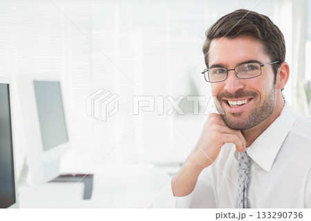 Businessman smiling and resting chin on hand at office desk with monitor, keyboard, copy space Businessman smiling and resting chin on hand at office desk with monitor, keyboard, copy space 133290736
