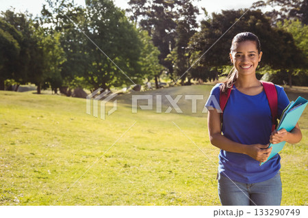 Female student standing on lawn holding notebooks and folders wearing red backpack, copy space 133290749