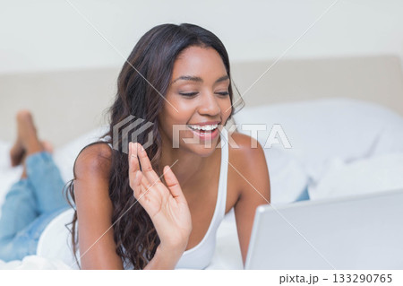 African American woman reclining on bed wearing white tank top, blue jeans waving at silver laptop 133290765