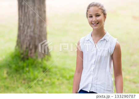 Smiling female child standing next to large tree trunk outdoors on green grass field, copy space Smiling female child standing next to large tree trunk outdoors on green grass field, copy space 133290787