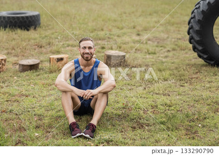 Adult male fitness enthusiast in blue top sitting, smiling on field with tractor tires and stumps Adult male fitness enthusiast in blue top sitting, smiling on field with tractor tires and stumps 133290790