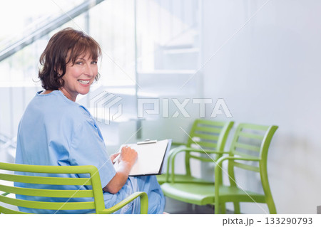 Female nurse sitting on green chair in waiting room writing on clipboard smiling back, copy space 133290793