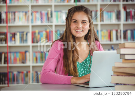 Teen female student sitting at round table in library reading using laptop and textbooks Teen female student sitting at round table in library reading using laptop and textbooks 133290794