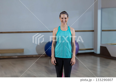 Woman smiling in sportswear while standing in studio with exercise balls, barre, mirror Woman smiling in sportswear while standing in studio with exercise balls, barre, mirror 133290807