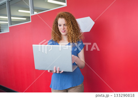 Twenty-something woman standing in red corridor holding silver laptop with white arrow sign 133290818